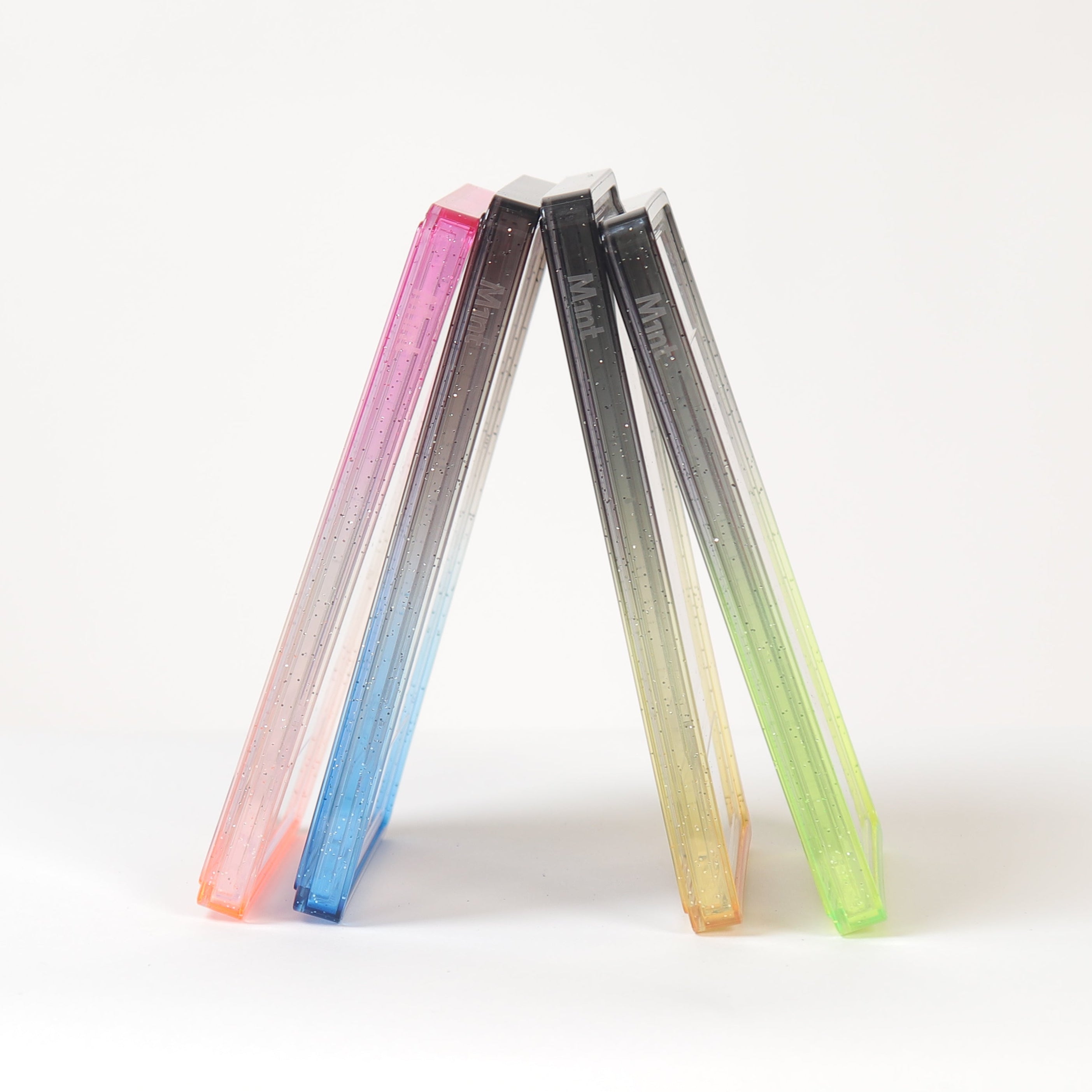 Three slim rectangular translucent items standing upright in a V shape on a white surface against a white background. The left item fades from pink to blue, the middle from black to gray, and the right from yellow to green.
